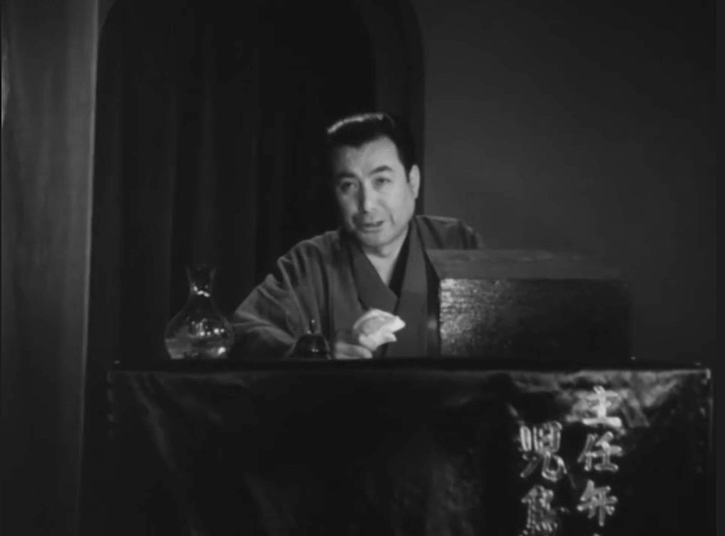 Archival still: a Japanese silent-film narrator in traditional dress at a narrators table beside the screen